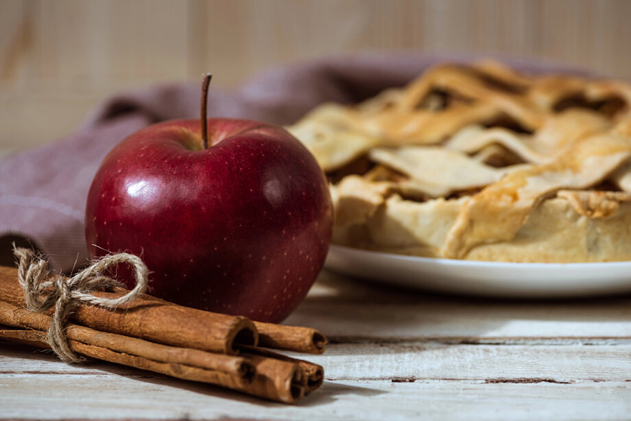 Im Hintergrund ein Apfelkuchen im Vordergrund ein Apfel und Zimtstangen